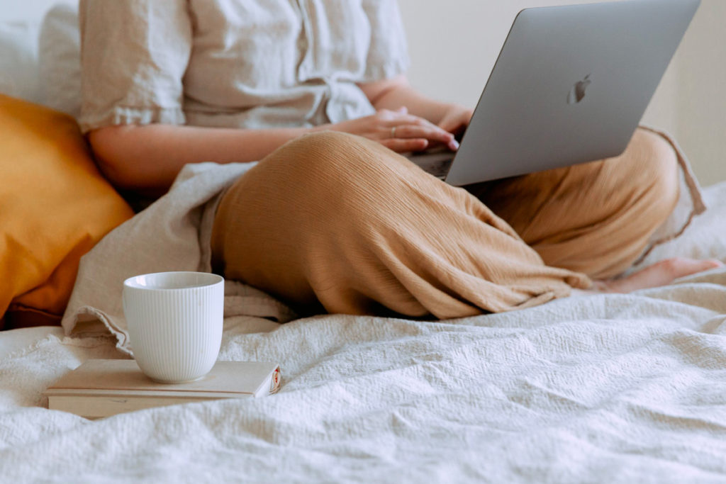 femme assise en tailleur sur un lit avec un tasse de thé et un macbook